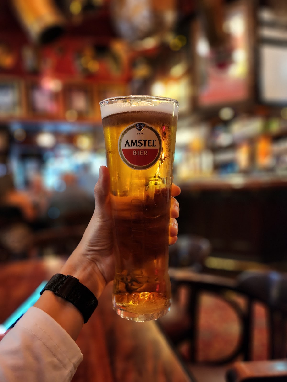 A hand holding up a tall glass of lager in front of the blurred, warmly lit interior of the pub.