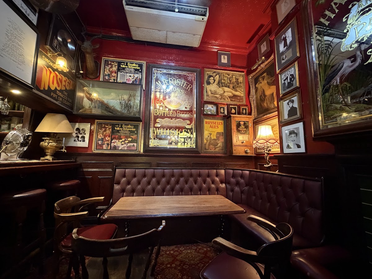 A corner of the bar inside: a buttoned oxblood leather banquette and bentwood chairs around a wooden table, under a bright red ceiling and walls hung with gilt-framed pictures, old brewery mirrors and enamel tobacco advertisements.