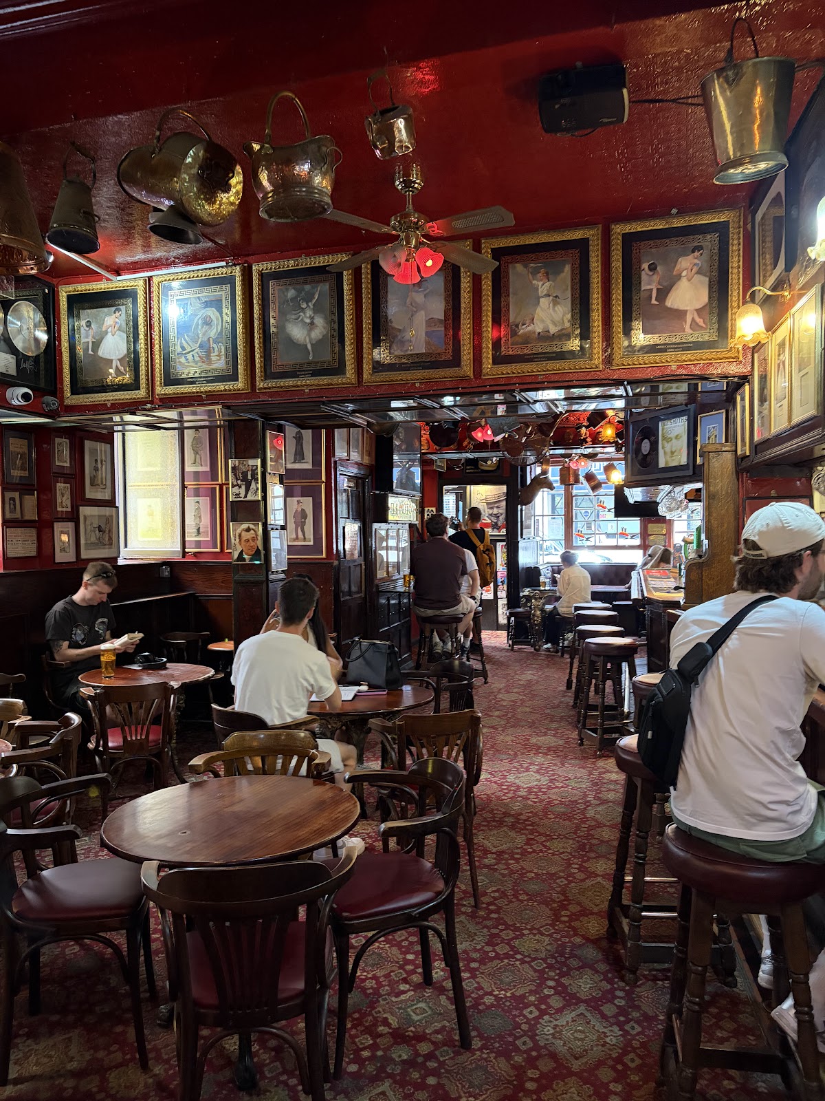 The main room looking towards the street door: patterned red carpet, round wooden tables and bar stools, customers drinking, with copper pans hung from the red ceiling and framed ballet paintings running along the top of the walls.