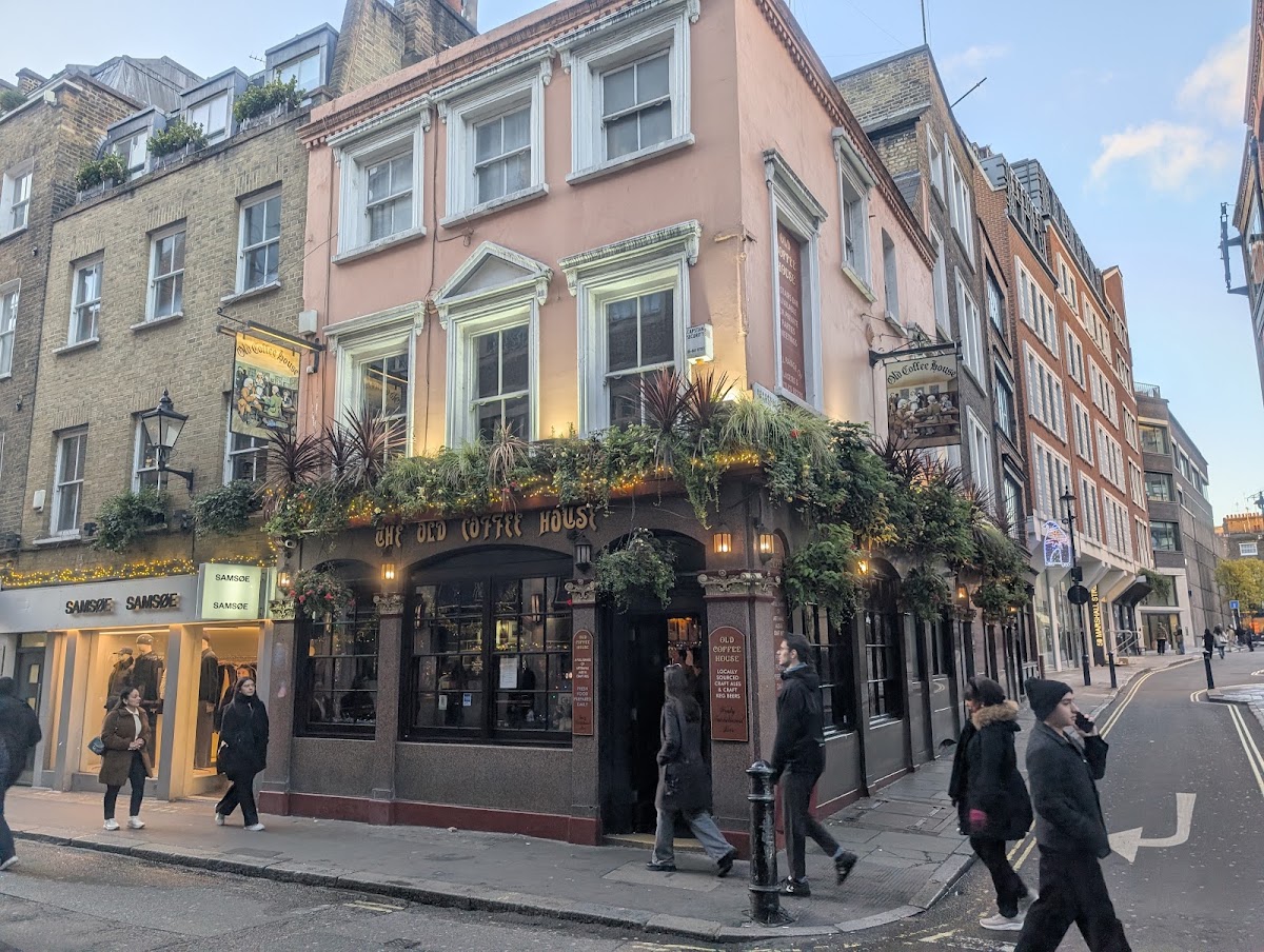 The Old Coffee House on a street corner: a pink stucco upper storey over a dark red painted ground floor, gold lettering reading THE OLD COFFEE HOUSE across the fascia, window boxes and a hanging pub sign, with people walking past on the pavement.