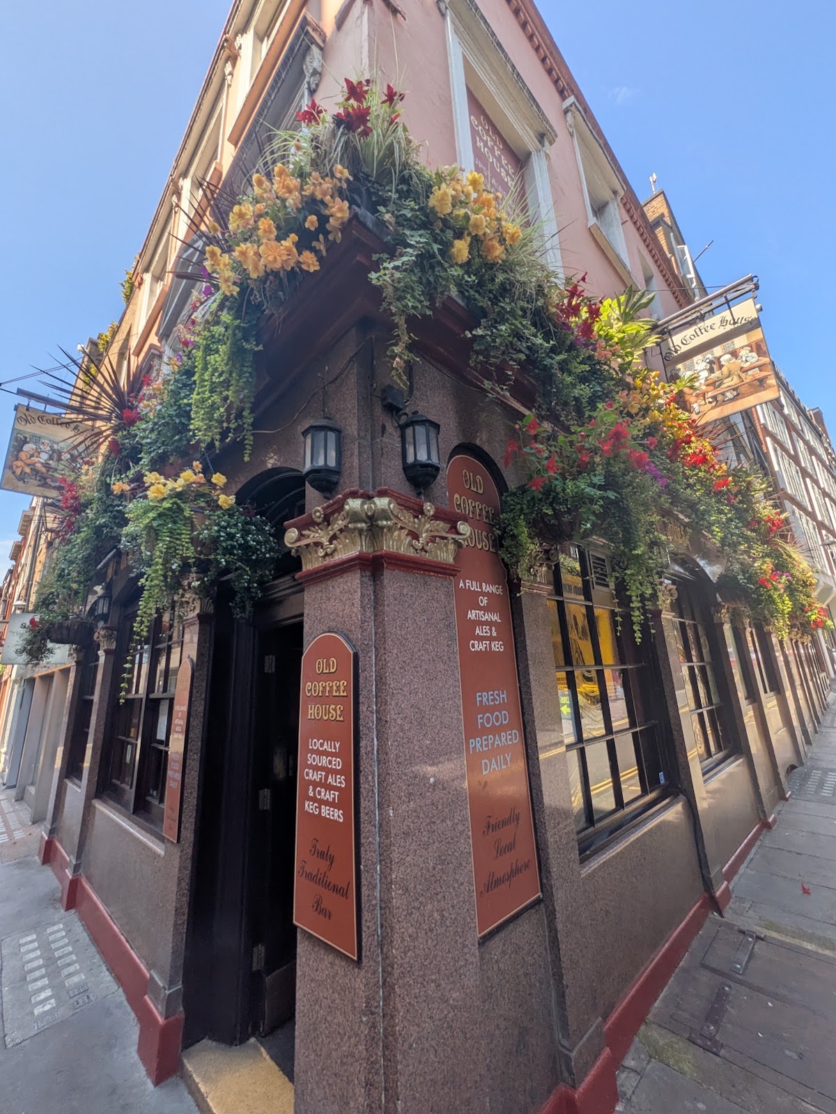 The pub's corner entrance from below: hanging baskets of red and yellow flowers over terracotta signboards with gold lettering reading OLD COFFEE HOUSE, LOCALLY SOURCED CRAFT ALES AND CRAFT KEG BEERS, FRESH FOOD PREPARED DAILY.